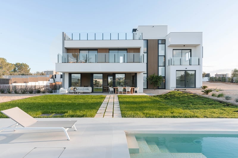 Moderne weiße zweistöckige Villa mit türkisfarbenem Pool, Sonnenliege und grünem Rasen unter klarem Himmel.