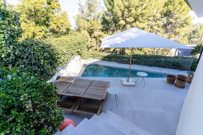 Terrasse en pierre avec piscine privée, chaises longues en bois et parasol blanc entourée de pins.