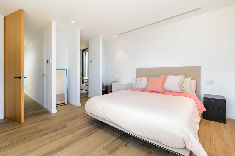 Chambre moderne et lumineuse avec un grand lit, des coussins roses, un sol en bois clair et des murs blancs.