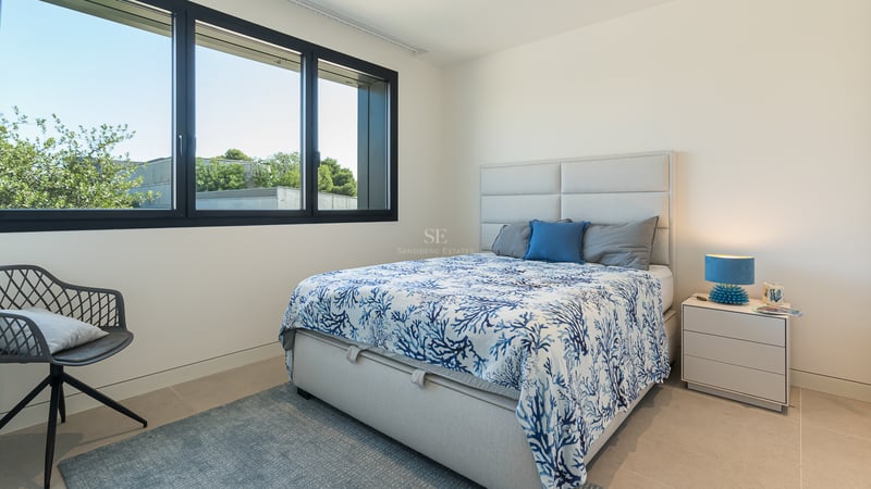 Chambre contemporaine avec grande fenêtre, lit aux motifs bleus, table de chevet blanche et chaise design.