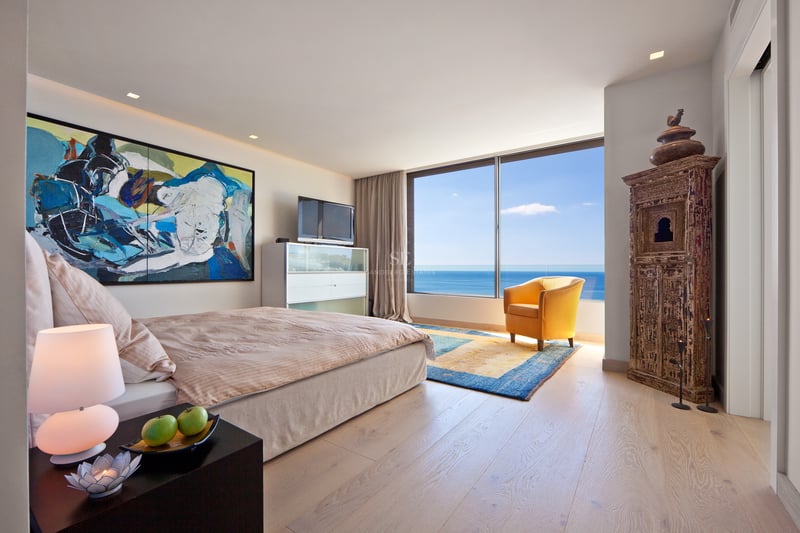 Chambre moderne avec vue sur mer, grand lit, fauteuil jaune et armoire en bois sculpté.
