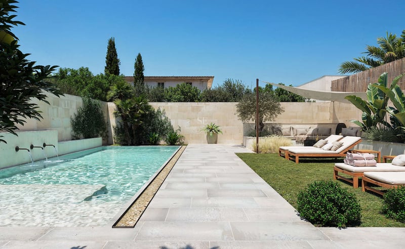 Piscine rectangulaire moderne avec jets d'eau, terrasse en pierre, bains de soleil en bois et plantes exotiques.