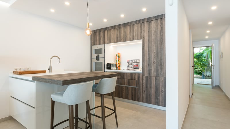 Cuisine contemporaine avec placards en bois sombre, îlot blanc et vue sur l'entrée du jardin au fond du couloir.