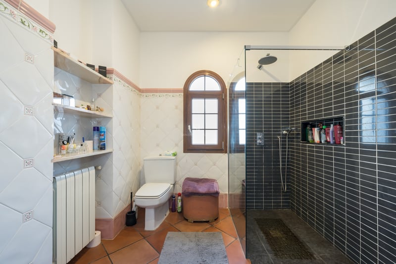 Salle de bain avec sol en terre cuite, carrelage décoratif, fenêtre cintrée en bois et douche moderne vitrée.