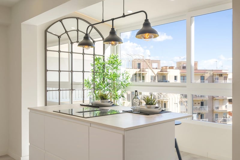 Îlot de cuisine blanc avec plaque à induction, suspensions industrielles et grandes fenêtres vue ville.