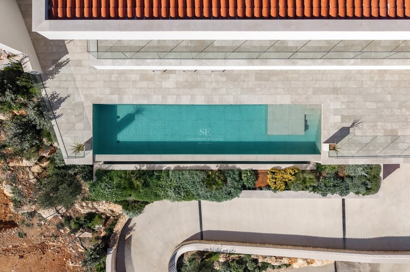 Vue plongeante d'une piscine turquoise, d'une terrasse en pierre, d'un toit en terre cuite et d'un jardin.
