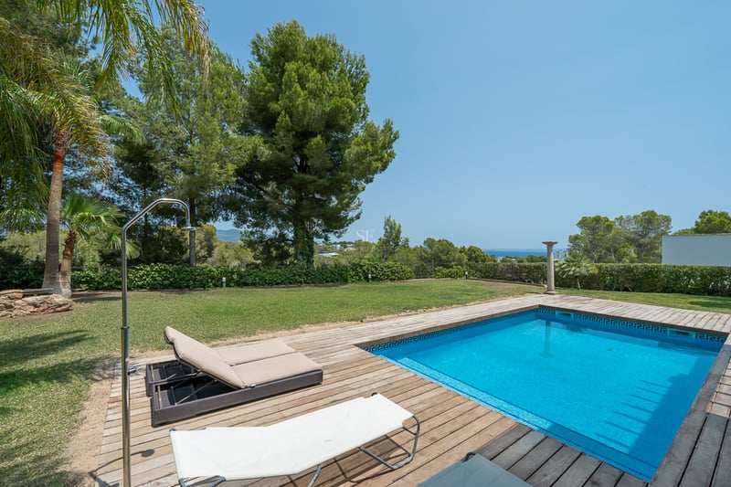 Piscine rectangulaire en mosaïque bleue entourée d'une terrasse en bois et d'une pelouse avec vue mer lointaine.