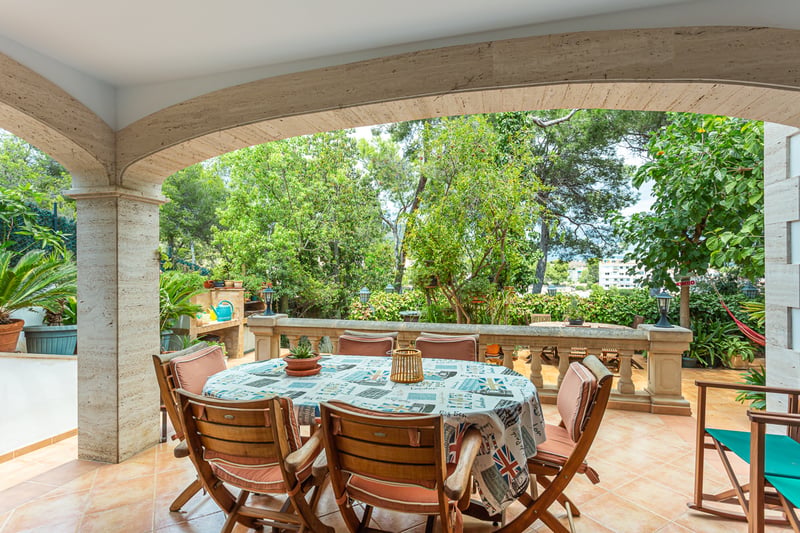 Terrasse en pierre couverte avec table ronde, chaises en bois et balustrade donnant sur un jardin verdoyant.