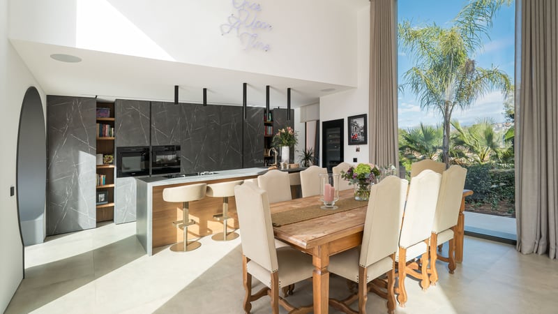 Salle à manger moderne avec table en bois, chaises rembourrées et cuisine en marbre sombre en arrière-plan.