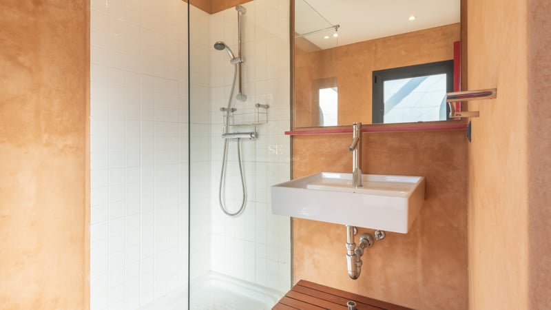 Salle de bain moderne avec douche en verre, carreaux blancs et murs texturés terre cuite avec vasque blanche.