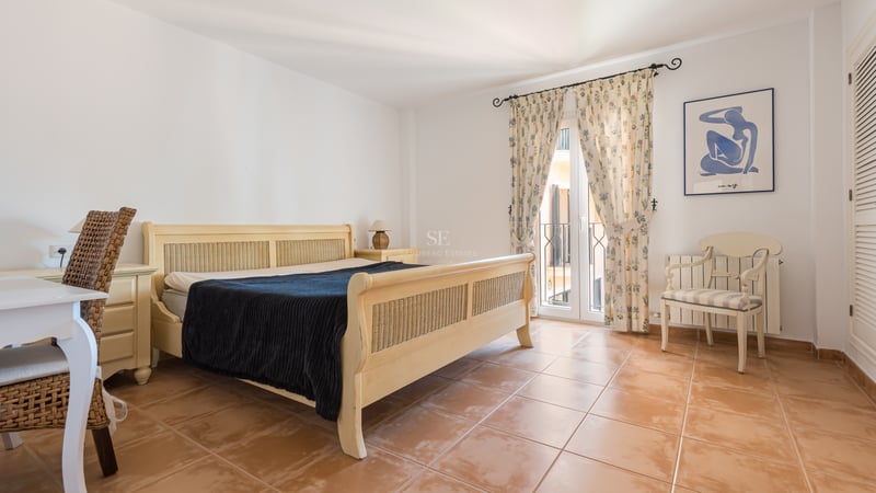 Chambre avec lit en osier, sol en terre cuite, bureau et balcon avec rideaux à motifs floraux.