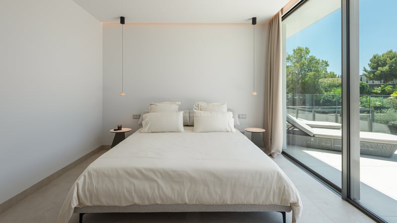 Chambre minimaliste avec grand lit, suspensions design et baies vitrées ouvrant sur une terrasse ensoleillée.