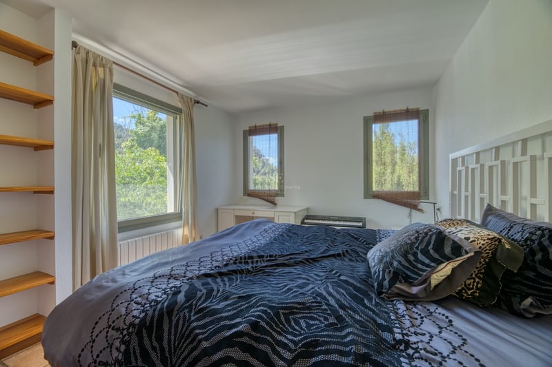 Chambre aux murs blancs, trois fenêtres donnant sur des arbres, lit avec coussins à motifs et étagères en bois.
