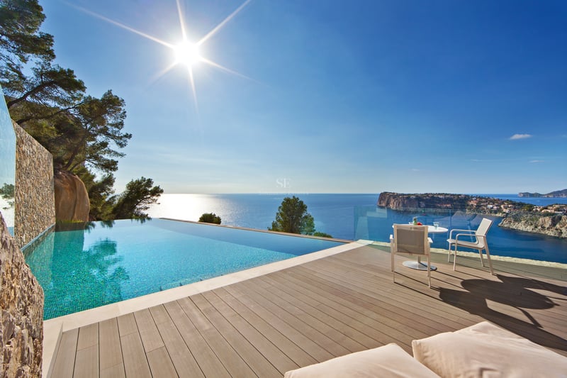 Piscine à débordement turquoise sur terrasse en bois avec vue sur mer bleue et côte rocheuse sous le soleil.