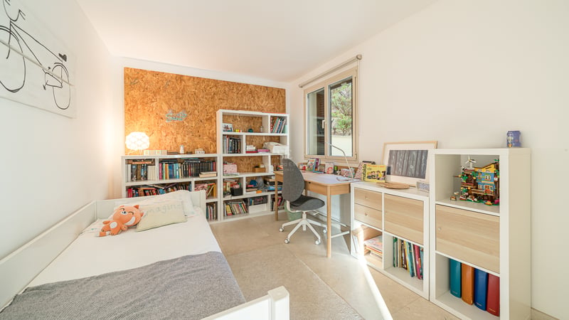 Chambre d'enfant avec lit blanc, bureau en bois et étagères remplies de livres sous une fenêtre ensoleillée.