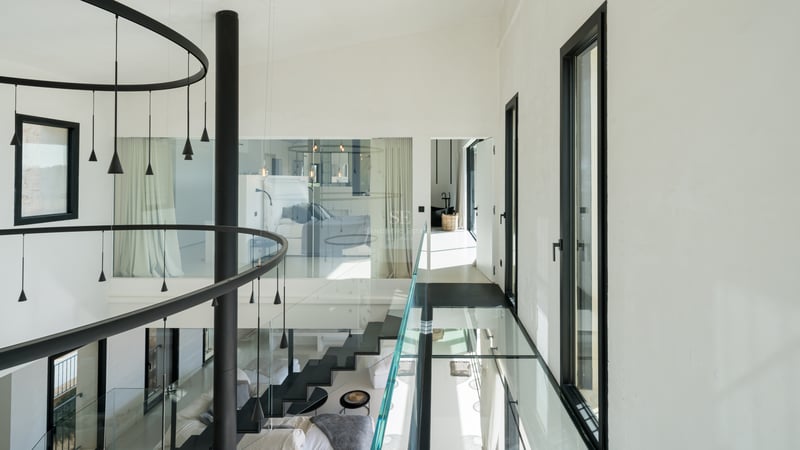 Couloir intérieur moderne avec passerelle en verre, escalier noir et grandes fenêtres dans une villa contemporaine.