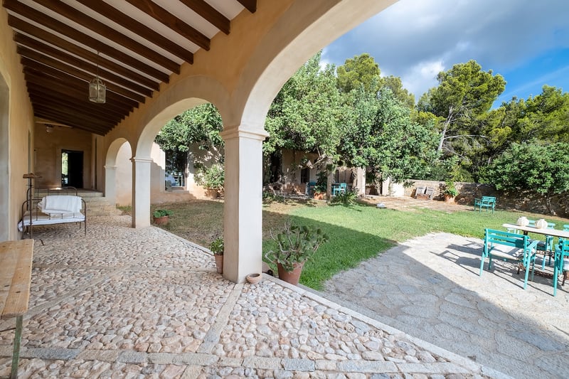 Porche traditionnel aux arches de pierre avec sol en pavés donnant sur un jardin ensoleillé avec meubles turquoise.