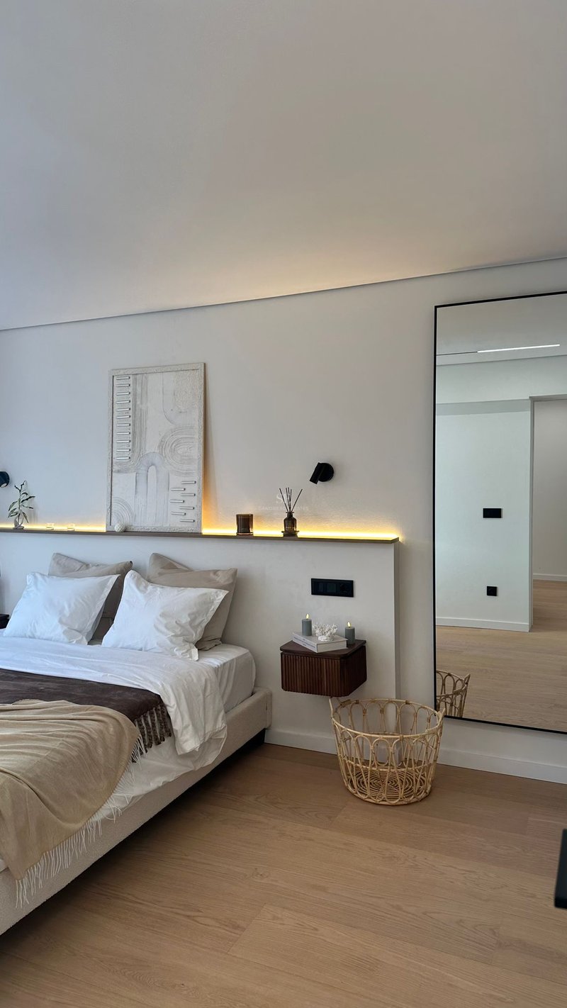 Chambre moderne avec lit blanc, rétroéclairage LED, œuvre d'art texturée et grand miroir sur parquet clair.