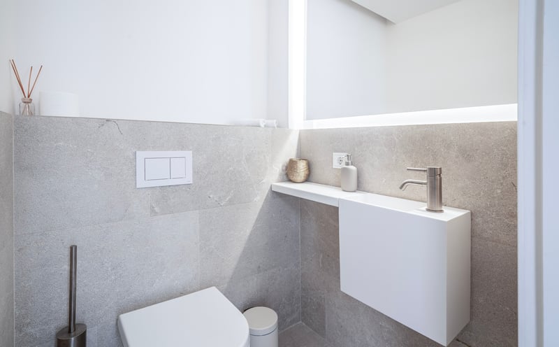 Salle de bain minimaliste avec WC blanc, petit lavabo suspendu et grand miroir rétroéclairé sur murs en pierre grise.
