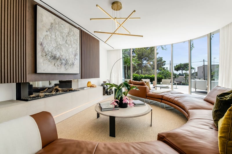 Salon moderne avec canapé courbé en cuir cognac, cheminée linéaire et grandes baies vitrées donnant sur un jardin.