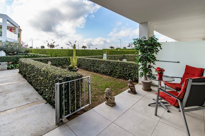 Terrasse blanche moderne avec deux chaises rouges et une table donnant sur un jardin privé aux haies taillées.