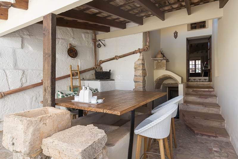 Rustic outdoor dining area with wooden table, stone walls, exposed beams, and stone steps leading to an interior.