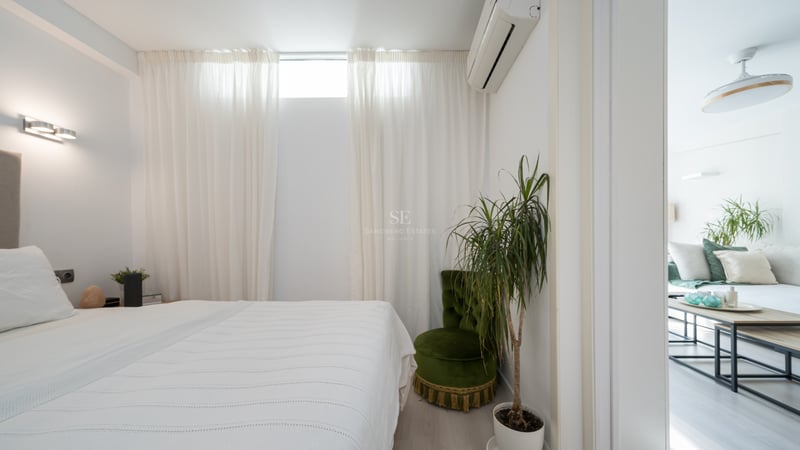 Chambre blanche épurée avec lit, fauteuil en velours vert, plante verte et vue sur le salon attenant.
