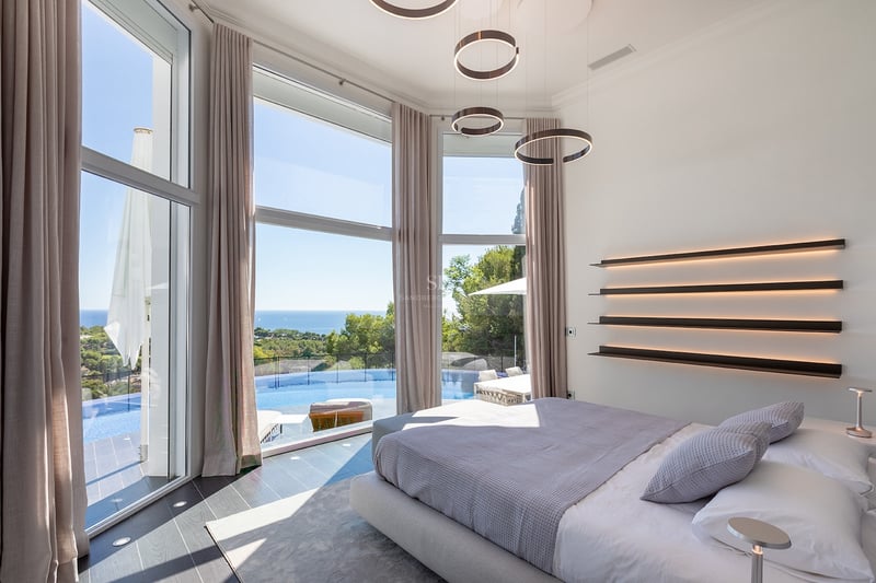 Chambre moderne avec de grandes baies vitrées donnant sur une piscine à débordement et la mer Méditerranée.