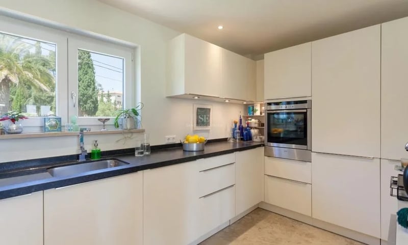 Cuisine contemporaine avec placards blancs sans poignées, plans de travail en granit noir et fenêtre sur le jardin.
