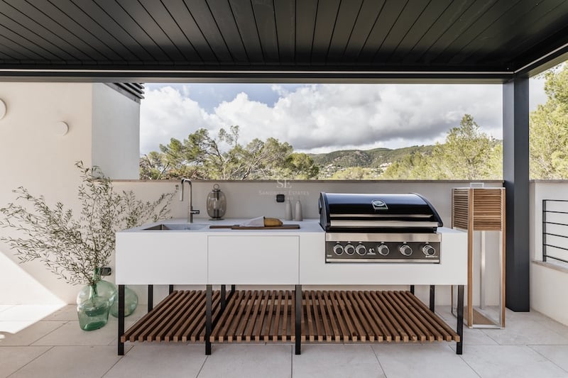 Cuisine d'été moderne avec barbecue professionnel, évier et meubles blancs sur une terrasse surplombant les montagnes.