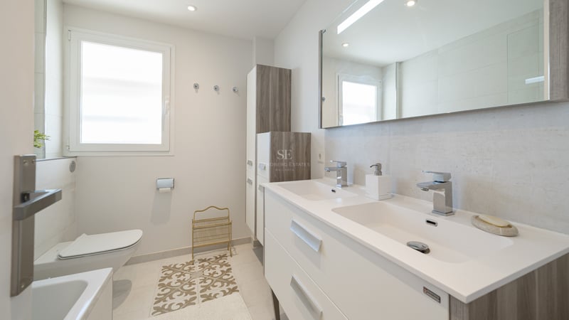 Salle de bain moderne lumineuse avec double vasque blanche, grand miroir, WC suspendu et lumière naturelle.
