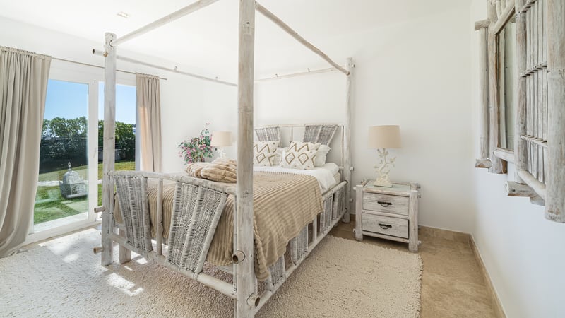 Chambre lumineuse avec lit à baldaquin en bambou blanc, textures tissées et accès direct au jardin.