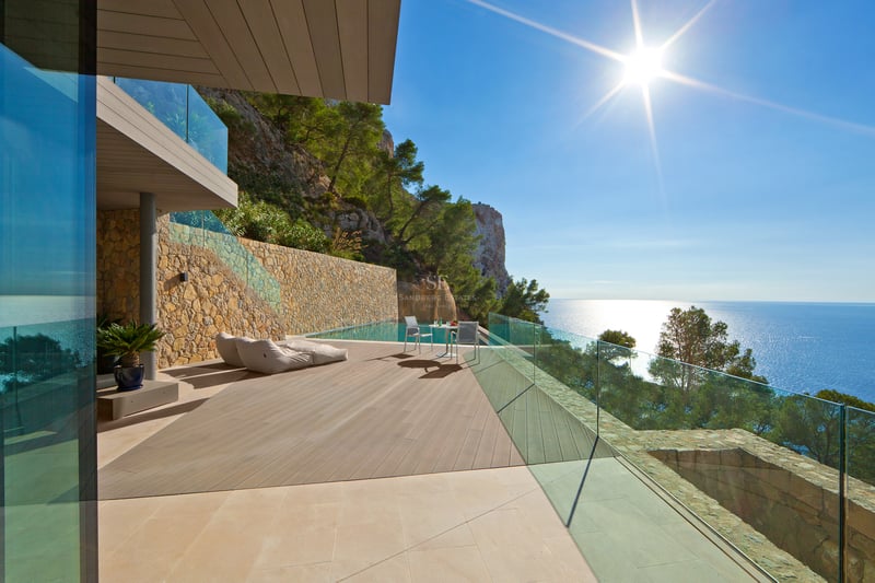 Terrasse de villa moderne avec piscine à débordement, deck en bois, garde-corps en verre et vue panoramique sur la mer.