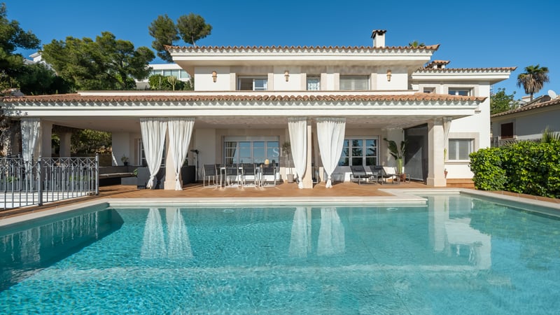 Vue d'une villa de luxe blanche avec grande piscine, terrasse couverte et rideaux blancs sous un ciel bleu.