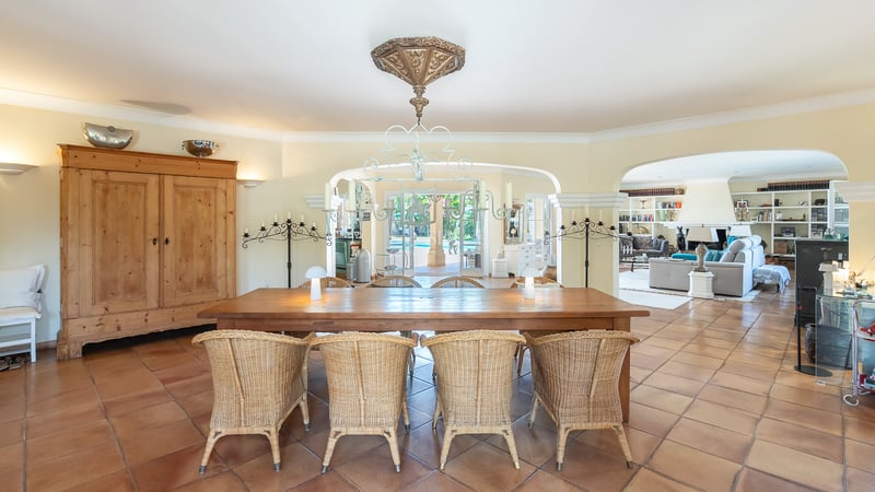 Spacious dining area with a long wooden table, wicker chairs, terracotta flooring, and arched views to the pool.