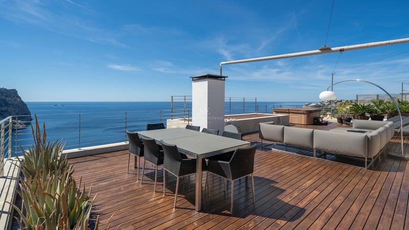 Terrasse moderne avec parquet en bois, table à manger, coin salon et garde-corps en verre surplombant la mer.