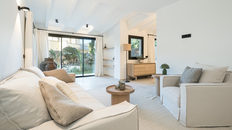 Salon lumineux avec plafonds à poutres blanches, canapés crème et baies vitrées ouvrant sur un jardin luxuriant.