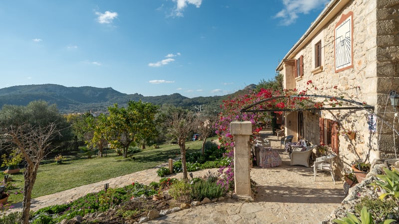 Terrasse en pierre avec mobilier extérieur, bougainvilliers roses et vue sur les montagnes sous un ciel bleu.