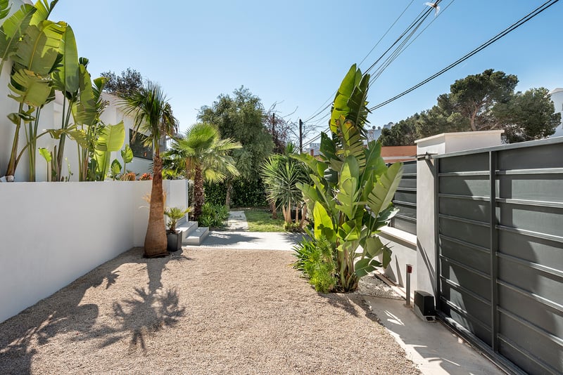 Portail coulissant gris foncé, mur blanc et zone de gravier entourée de plantes tropicales sous un ciel bleu.