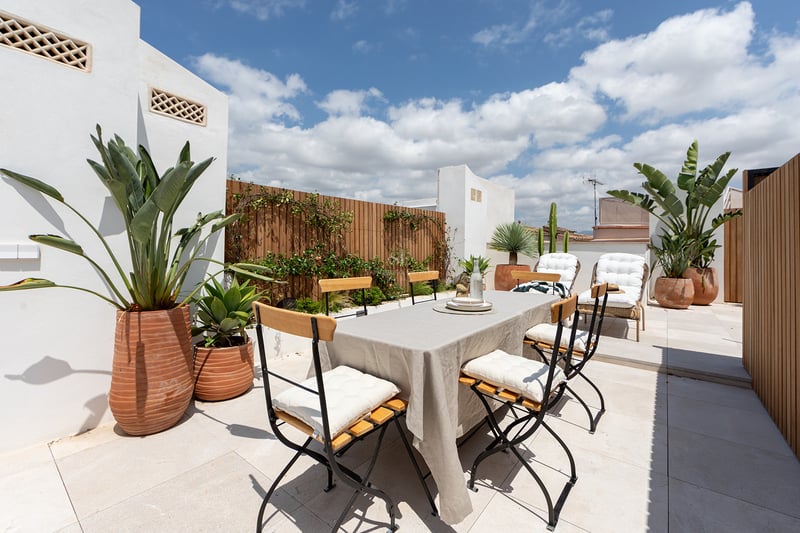 Terraza privada con mesa de comedor, mantelería de lino, valla de madera y plantas tropicales bajo cielo azul.