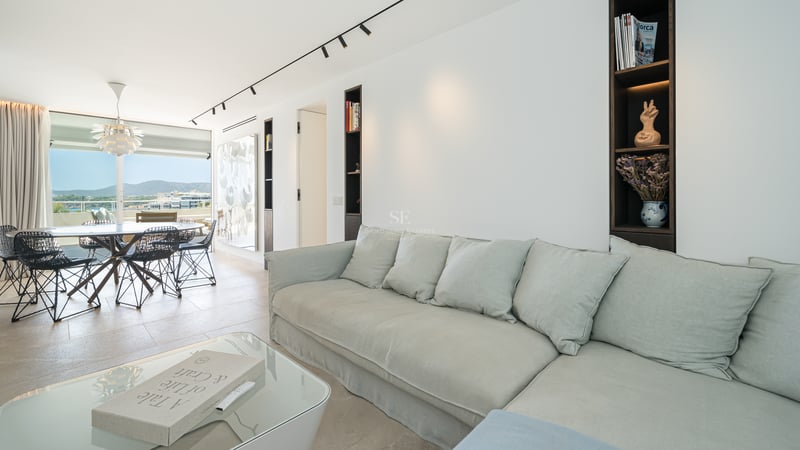 Salon lumineux avec un grand canapé gris clair, table basse en verre et salle à manger en arrière-plan avec vue sur la mer.