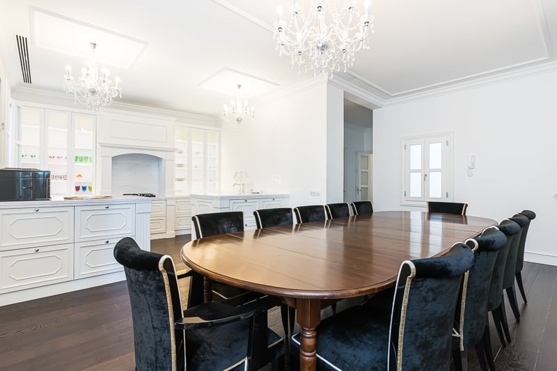 Grande table ovale en bois avec chaises en velours noir sous des lustres en cristal dans une pièce blanche.