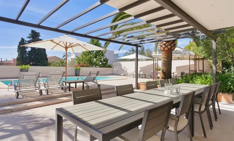 Pergola moderne grise abritant une table à manger extérieure à côté d'une piscine avec chaises longues et palmiers.