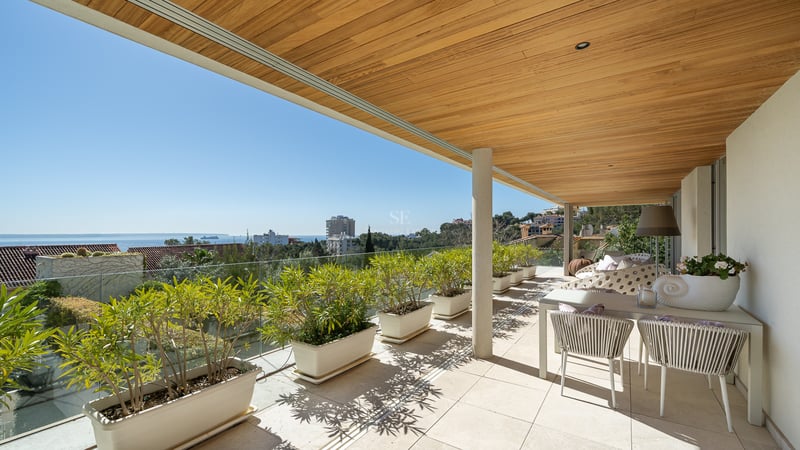 Terrasse couverte avec plafond en bois, garde-corps en verre et jardinières blanches offrant une vue sur la ville.