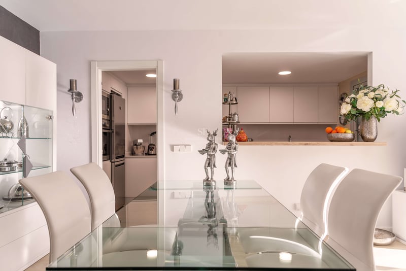Salle à manger contemporaine avec table en verre, chaises blanches et vue sur une cuisine blanche moderne.
