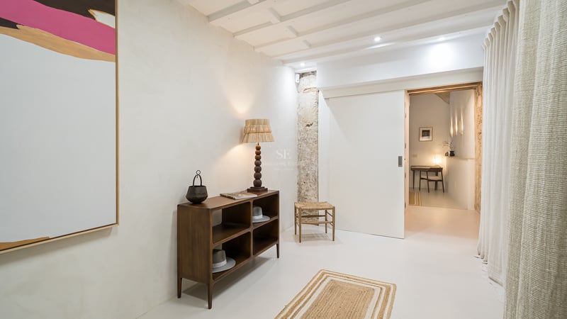 Couloir moderne avec murs en plâtre blanc, console en bois, lampe rustique et vue sur un bureau.