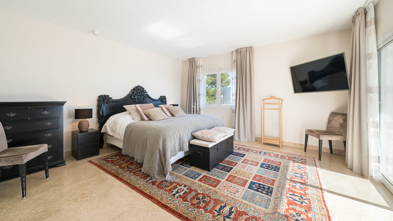 Chambre principale lumineuse avec tête de lit noire ornée, tapis à motifs et sol en travertin.