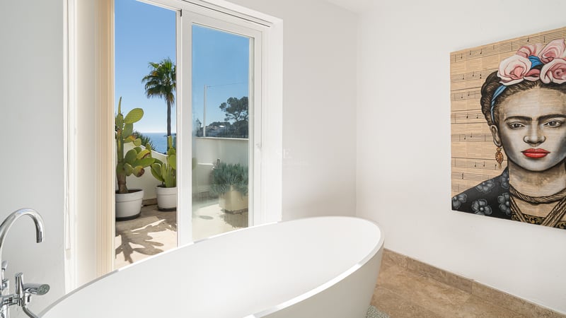 Salle de bain moderne avec baignoire blanche, portrait de Frida Kahlo et terrasse avec vue sur la mer.
