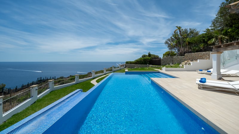 Piscine à débordement bleue surplombant la mer avec transats sur une terrasse en pierre sous un ciel bleu.