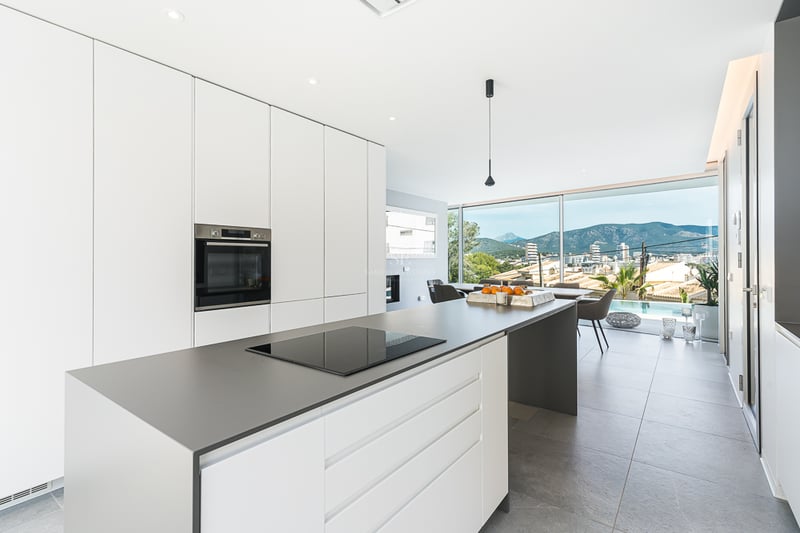 Cuisine blanche lumineuse avec îlot gris, plaque à induction et grandes baies vitrées avec vue sur la montagne.